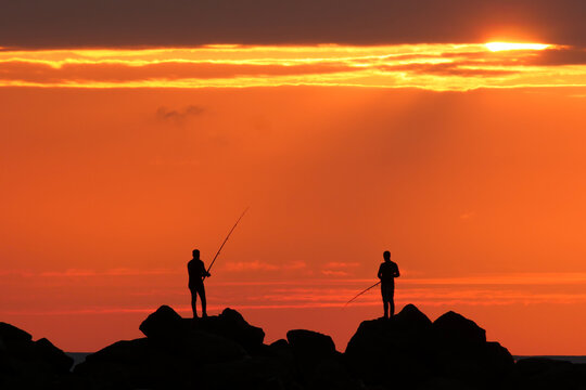 People fishing with a fishing rod at sunset. Impressive image of red sky and sun. - Powered by Adobe