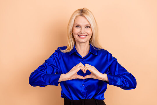 Smiling middle aged woman in blue shirt makes heart sign with hands on beige background - Powered by Adobe