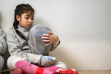 A girl in fencing gear is sitting on the sidelines, feeling disappoint for the unsuccessful game