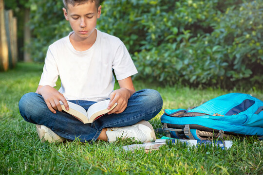 Schoolboy teen (teenager) seating and reading in the city park. Young smiling boy studying on the grass, near pupil (student) is backpack. Sunny day. Education concept.
