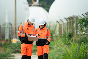 Two refinery workers discuss data on laptop, Backgroud of large storage tank, engineer communicate using walkie-talkie, industrial site operation