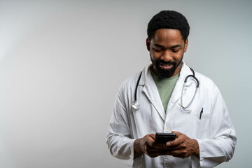 African american doctor smiling while using smartphone on isolated background, sending messages or browsing social media, relaxed mood and modern lifestyle