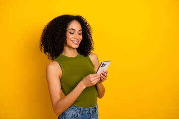 Charming young woman in stylish outfit using smartphone against vibrant yellow background...