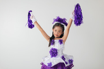 Little cheerleader girl in purple uniform with pom-poms, smiling joyfully. White studio background highlights her enthusiasm and team spirit. cheerleader kid
