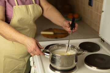 Senior woman's hands stirring sweet cream ghee with a metal spoon with holes in a saucepan on the stove