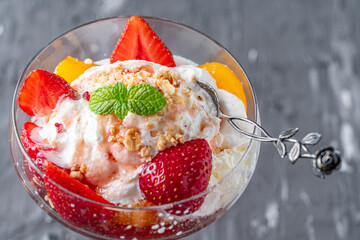 Ice cream with whipped cream, strawberries and peach in a glass tall glass on a gray background....