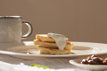 Pancakes with cream and jam on white plate with cup of coffee during breakfast, enjoying morning sunlight