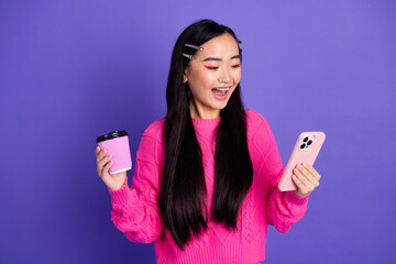 Excited young woman holding a coffee cup and phone against a vibrant purple background, expressing...