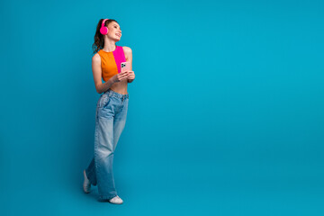 Cheerful young woman listening to music with pink headphones holding a smartphone on a stylish blue...