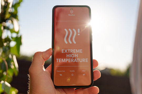 Smartphone screen showing extreme high temperature warning alert, hand holding phone under bright sunny sky and hot day