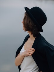 Young woman wearing bowler hat and holding blazer posing by water