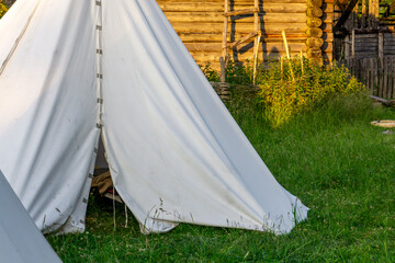 White canvas historical tents set up on green grass near wooden log structures. A scene from a...