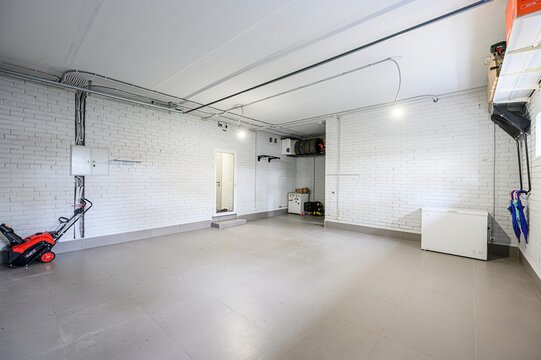 Clean, well-lit garage with white brick walls and gray tiled floor. A snowblower is near an electrical panel. A chest freezer sits against a wall