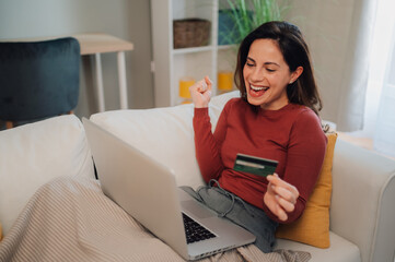 Excited woman celebrating online shopping success with credit card and laptop on sofa