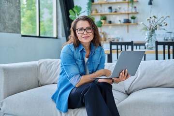 Obraz premium Portrait of middle aged woman with laptop sitting on sofa at home