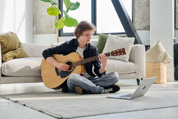 Teenage boy with acoustic guitar and laptop, having online music lesson