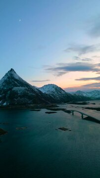 Ramberg auf den Lofoten in Norwegen bei Sonnenuntergang im Winter, Stra&szlig;e f&uuml;hrt &uuml;ber Br&uuml;cke zum schneebedeckten Berg, weiches Licht &uuml;ber K&uuml;ste, Meer und felsiger arktischer Landschaft