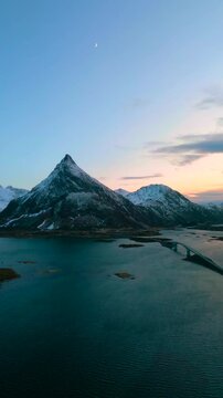 Ramberg auf den Lofoten in Norwegen bei Sonnenuntergang im Winter, Stra&szlig;e f&uuml;hrt &uuml;ber Br&uuml;cke zum schneebedeckten Berg, weiches Licht &uuml;ber K&uuml;ste, Meer und felsiger arktischer Landschaft