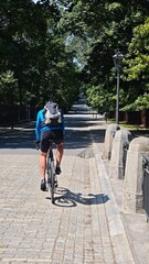 A cyclist riding alone on an empty park road surrounded by lush green trees, wearing a blue jacket and backpack