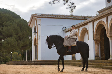 Young horsewoman riding a black Spanish thoroughbred horse next to an old hermitage. She has a...
