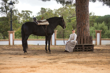 Young woman rider sitting on a wooden bench posing next to her black Spanish thoroughbred horse,...