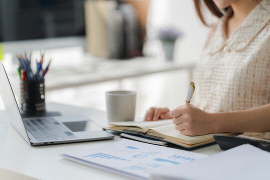 Focused Work: A woman is deeply engrossed in her work, taking notes in a notebook at her tidy desk with computer and financial reports nearby, capturing the essence of professional concentration. - Powered by Adobe