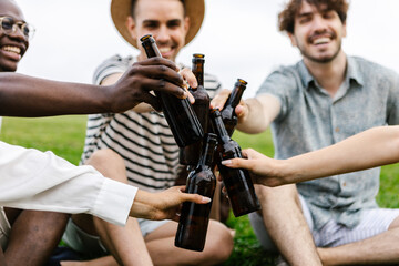 Diverse friends cheering with beer bottles while enjoying summer picnic at city park