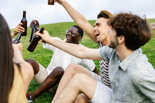 Young group of diverse friends having fun drinking beer during summer vacation, relaxing together sitting on grass at city park - Powered by Adobe