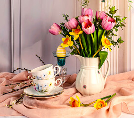 Spring still life with beautiful bouquet of pink tulip flowers, yellow daffodils in a white vintage jug, two cups of coffee, blooming green branches on a pastel light background