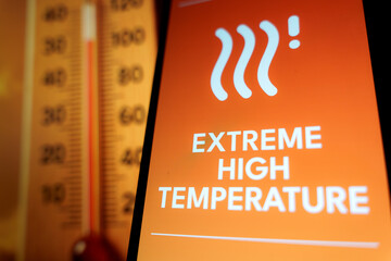 Smartphone screen showing extremely hot weather warning with thermometer in background, symbolizing heatwave.