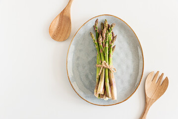 Fresh asparagus tied bundle on plate and wooden utensils on white table. Top view