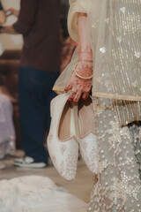 Bridal Elegance: A bride holds her embroidered shoes, adorned with henna and a gold bangle,...
