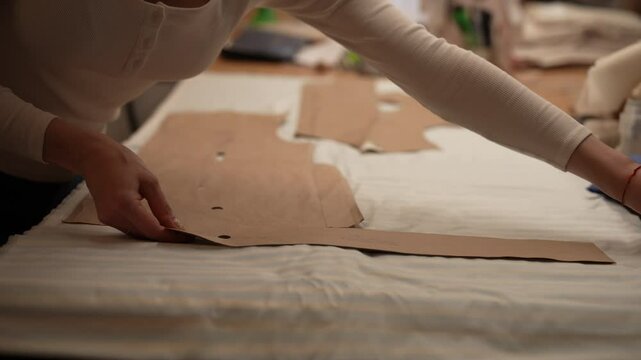 Professional tailor organizing cardboard sewing patterns on white striped fabric, readying material for garment construction in dressmaking workspace, close-up. Concept of sewing manufacturing.