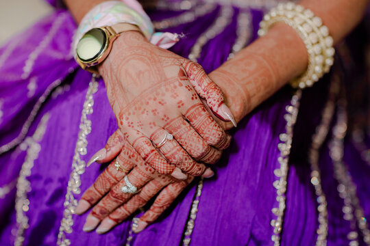 Hands adorned with intricate henna designs, jewelry, and watch. The purple fabric adds a vibrant contrast and festive atmosphere.