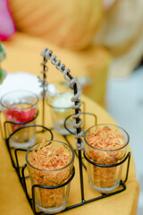 Close-up of glasses filled with colorful flower petals, arranged in a black metal stand on a vibrant orange surface. Decorative and celebratory.