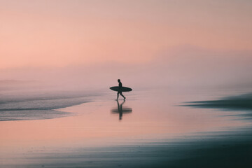 Silhouette of a surfer carrying a board on a misty beach at sunrise