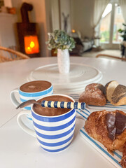 Blue and white striped mugs filled with hot chocolate, accompanied by assorted pastries on a cozy table, creating a warm and inviting breakfast atmosphere