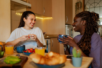 Happy lesbian couple eating fresh fruit and croissants, drinking orange juice and coffee or tea,...