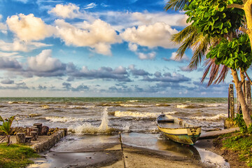 Small wooden boat on a tropical beach with palm trees and stormy ocean waves. Concept of travel, solitude, nature and adventure.