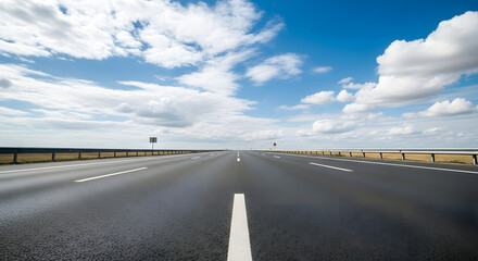 Fototapeta premium A long, straight asphalt road stretches towards the horizon under a bright blue sky with fluffy white clouds.