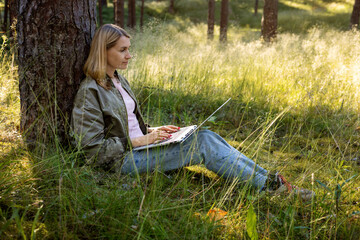 self-employed woman using laptop in the park. digital nomad, small business owner sitting against the tree and work remotely in nature. freelance remote working concept