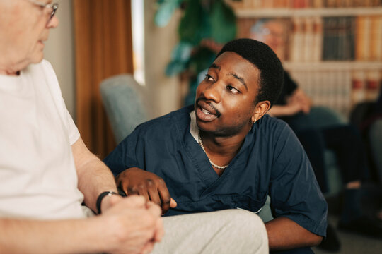 Young male nursing assistant consoling elderly man sitting in wheelchair at retirement home