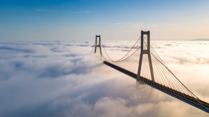 A long, modern cable-stayed bridge stretches into the distance, partially obscured by a thick blanket of low-lying clouds or fog, under a clear sky. This ethereal landscape evokes grandeur and mystery