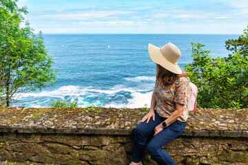 Tourist woman wearing hat sitting and contemplating the ocean views from Mount Urgull, San Sebastian, Spain.