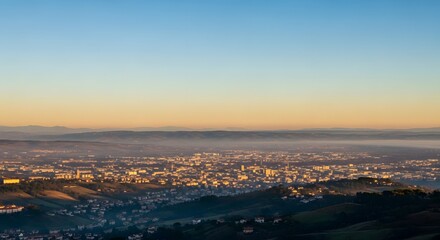 Aerial panorama captures a cityscape bathed in golden sunrise hues rising above rolling hills and subtle morning fog illuminating buildings and sky.