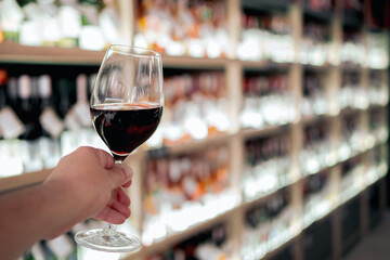 A hand holding a glass of red wine in a wine store with blurred bottles on shelves in the...