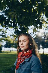 Naklejka premium lifestyle woman in modern boho-western denim street style wearing red bandana outdoors with natural light and film color tones, calm expression under green leaves