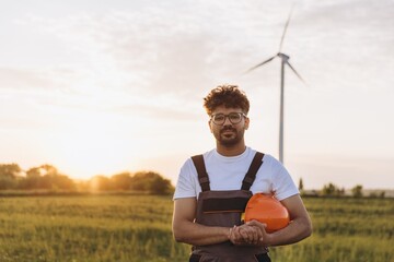 Young engineer posing in a field with a wind turbine at sunset