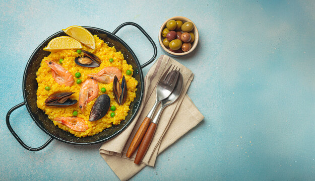 Seafood paella with rice, shrimp, mussels, peas, and lemon wedges served in a black traditional pan on a dark rustic background with olives and cutlery, Mediterranean meal, copy space