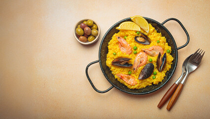 Seafood paella with rice, shrimp, mussels, peas, and lemon wedges served in a black traditional pan on a light beige background with olives and cutlery
